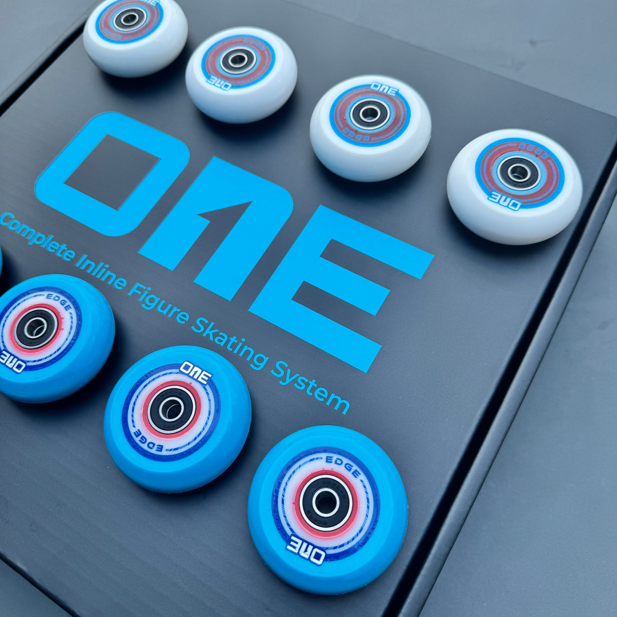 A set of inline figure skating wheels arranged on a dark gray box labeled 'ONE Complete Inline Figure Skating System.' The wheels are white and bright blue with multicolor center graphics, and the brand name 'ONE' is prominently visible on both the packaging and the wheels.