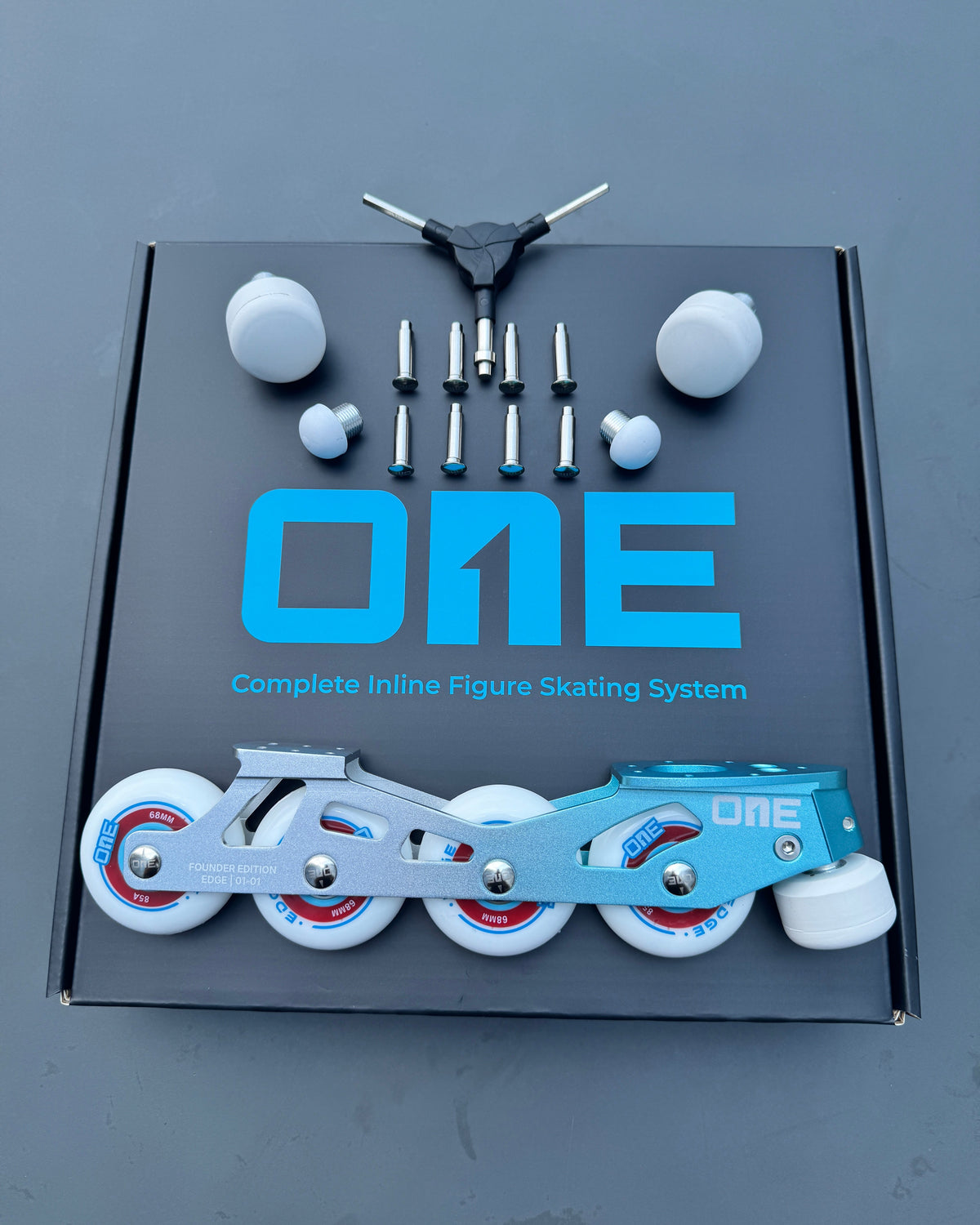 A complete inline figure skating system displayed on top of its black retail box. The set includes a light blue and silver inline skate with white wheels, a black trike, mounting hardware, and white wheel covers, all arranged neatly on a dark gray background.