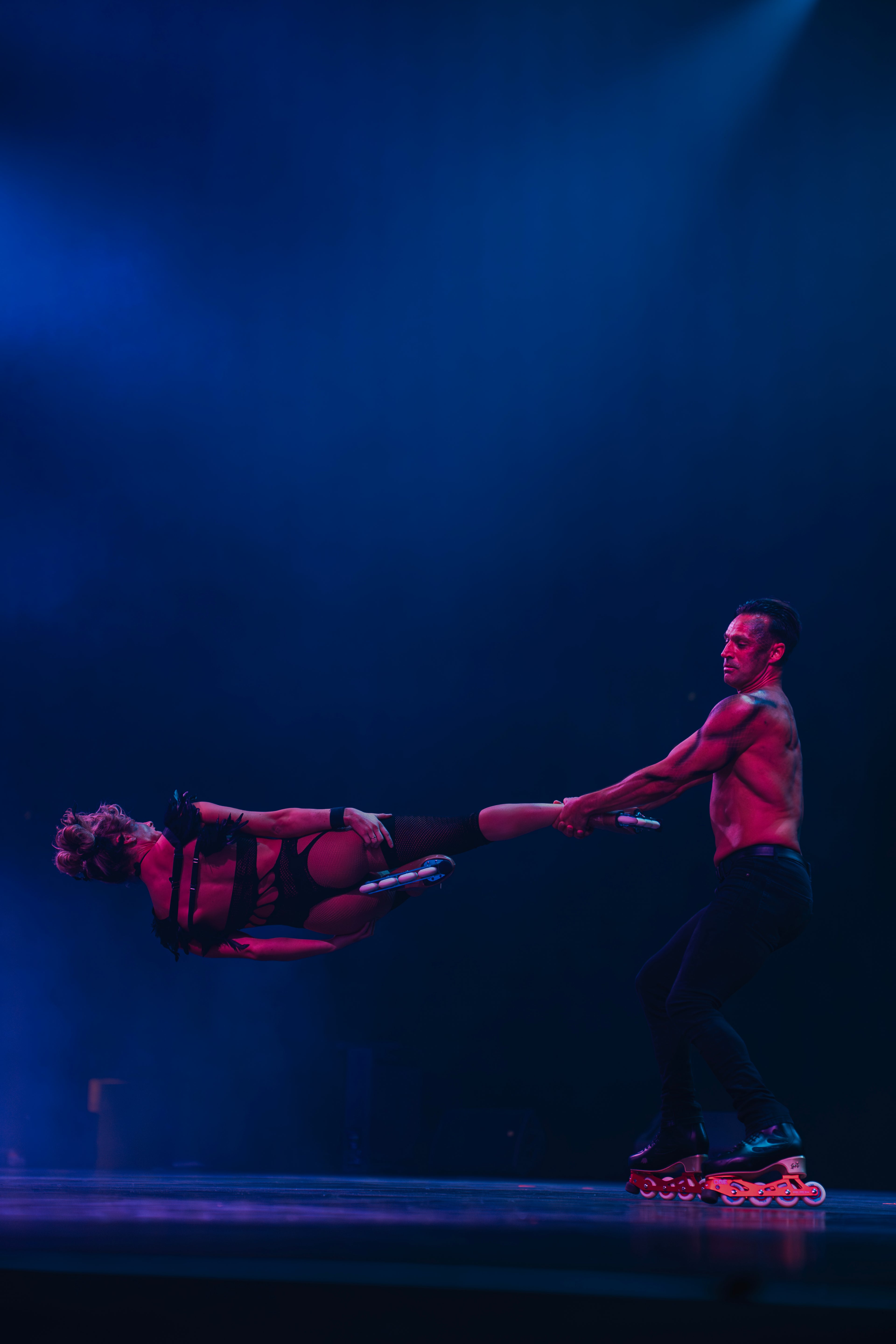 Rosie and Adam performing a headbanger skating trick for Little Freak show on a dark stage illuminated by dramatic blue spotlights